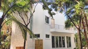 Ein modernes, zweistöckiges Haus mit Balkon, umgeben von Palmen und sandigem Boden.