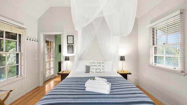 Un dormitorio con cama, ventana con persianas, mesita de noche con lámpara y puerta que da a otra habitación.