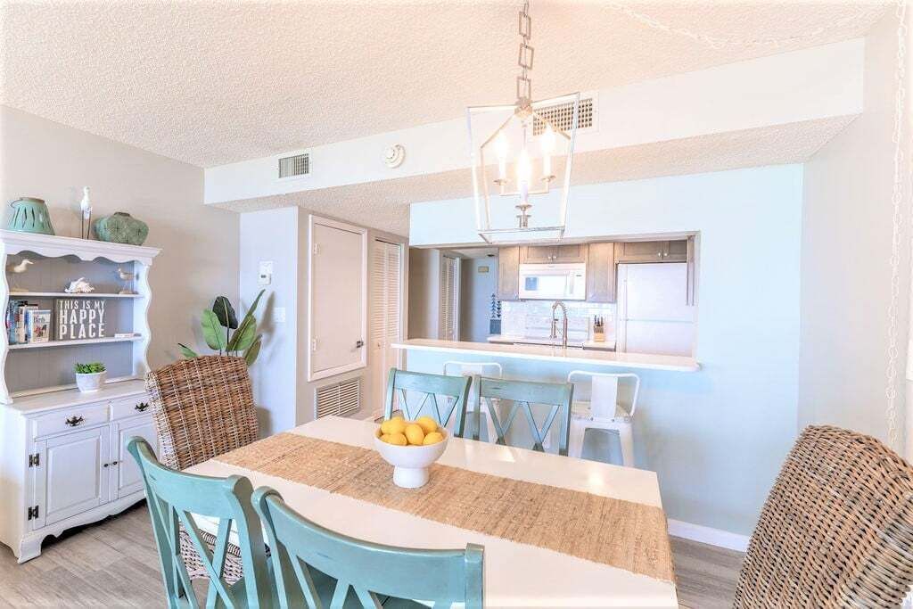 Un coin repas avec une table blanche, des chaises turquoise, un saladier de fruits et un luminaire suspendu.