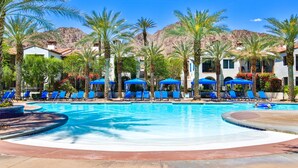 A swimming pool with blue lounge chairs and umbrellas, surrounded by palm trees and buildings.
