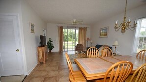 Uma área de refeições com uma grande mesa de madeira e cadeiras, um ventilador de teto e vista para o exterior através de uma porta de vidro.