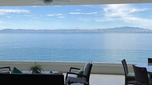 A modern interior with a ceiling fan, a sofa, and a table with chairs, overlooking a body of water and distant mountains.