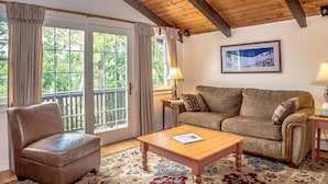 A cozy living room with a wooden ceiling, a brown leather sofa, a wooden coffee table, and a large window with curtains. - Abbott Brook Chalet A - Private Setting - 5 Mi  From Storyland (Intervale)