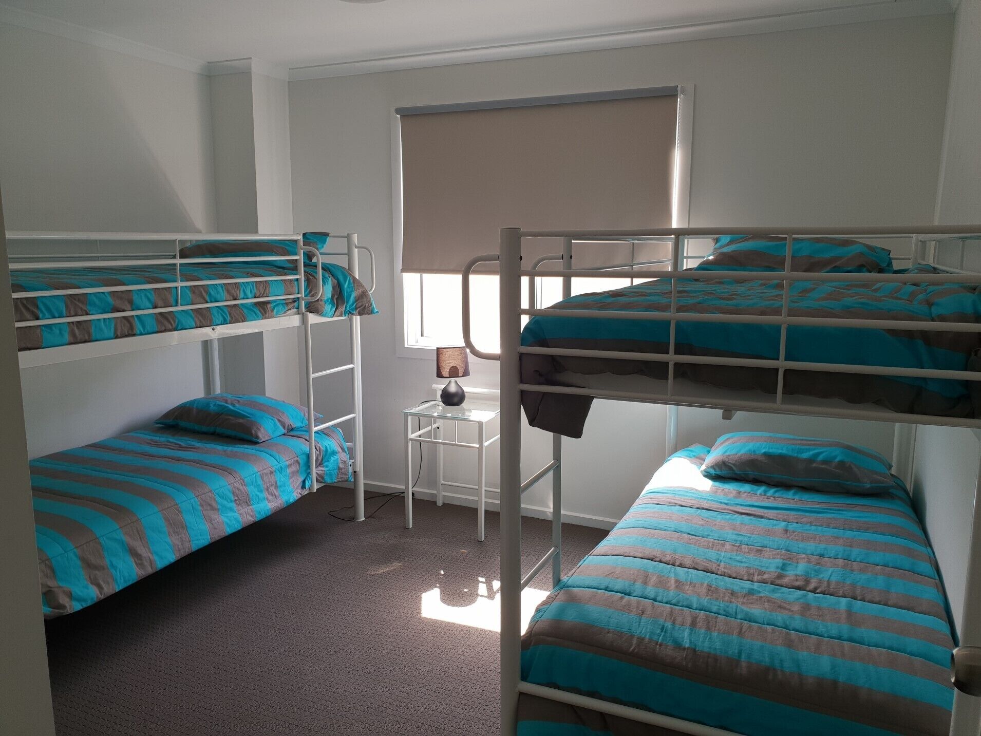 Une chambre avec un lit superposé, une couette à rayures, une petite table avec une lampe et une fenêtre avec des stores.