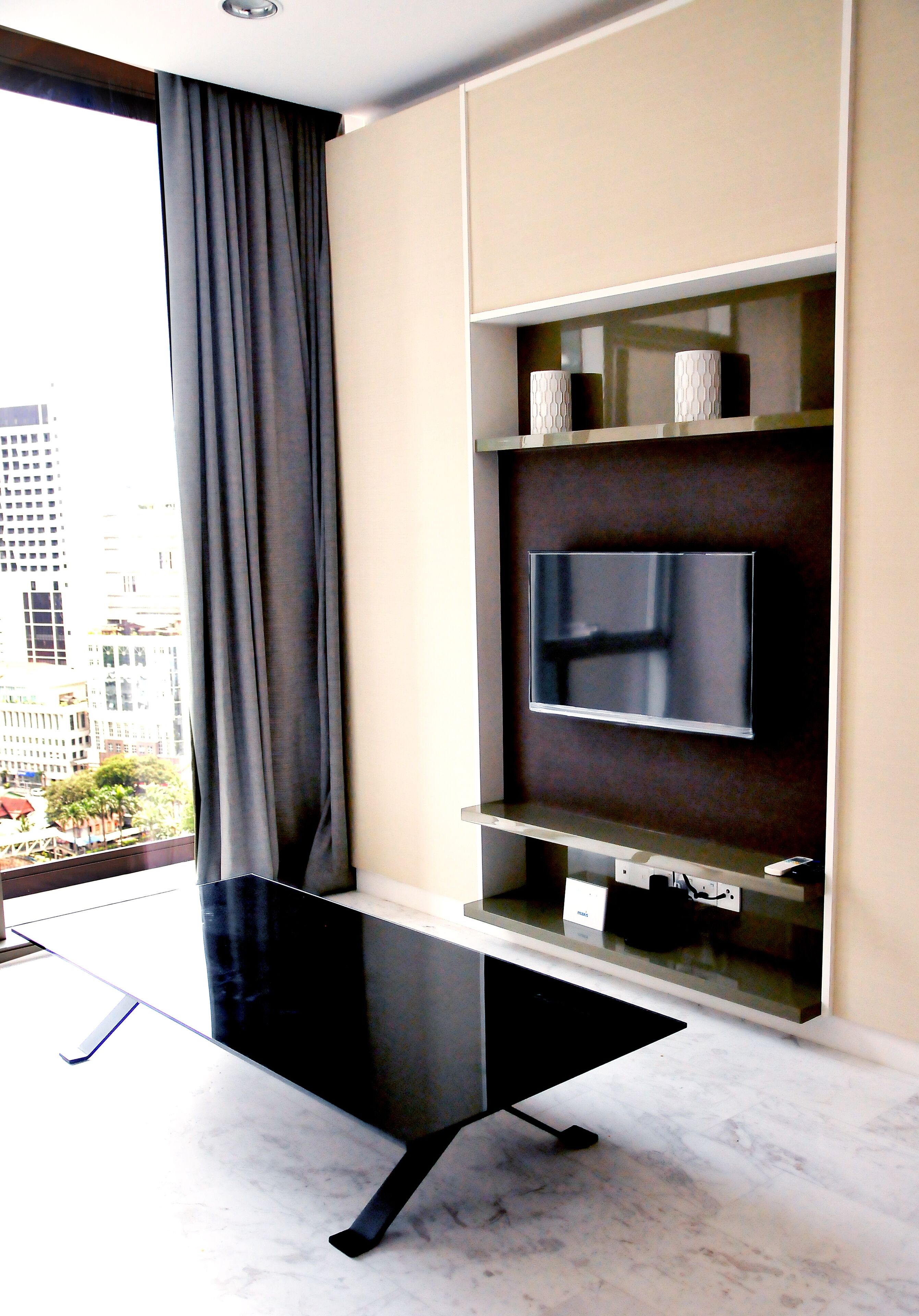 Un salon moderne avec un téléviseur à écran plat fixé au mur, une table basse noire au design épuré et une grande fenêtre donnant sur un paysage urbain.