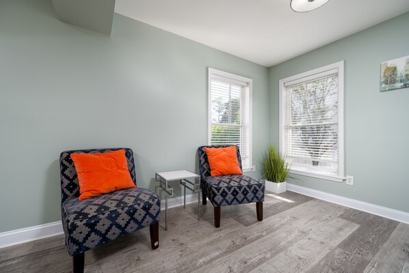 A room with two patterned armchairs with orange cushions, a small white side table, and a potted plant.