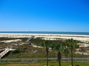 A beach with a wooden boardwalk leading to the shore, palm trees, and a fenced area with signs indicating pool hours. - Inn at Dauphin Island 309 Wonderful Unit with Expansive Views!! (Dauphin Island)