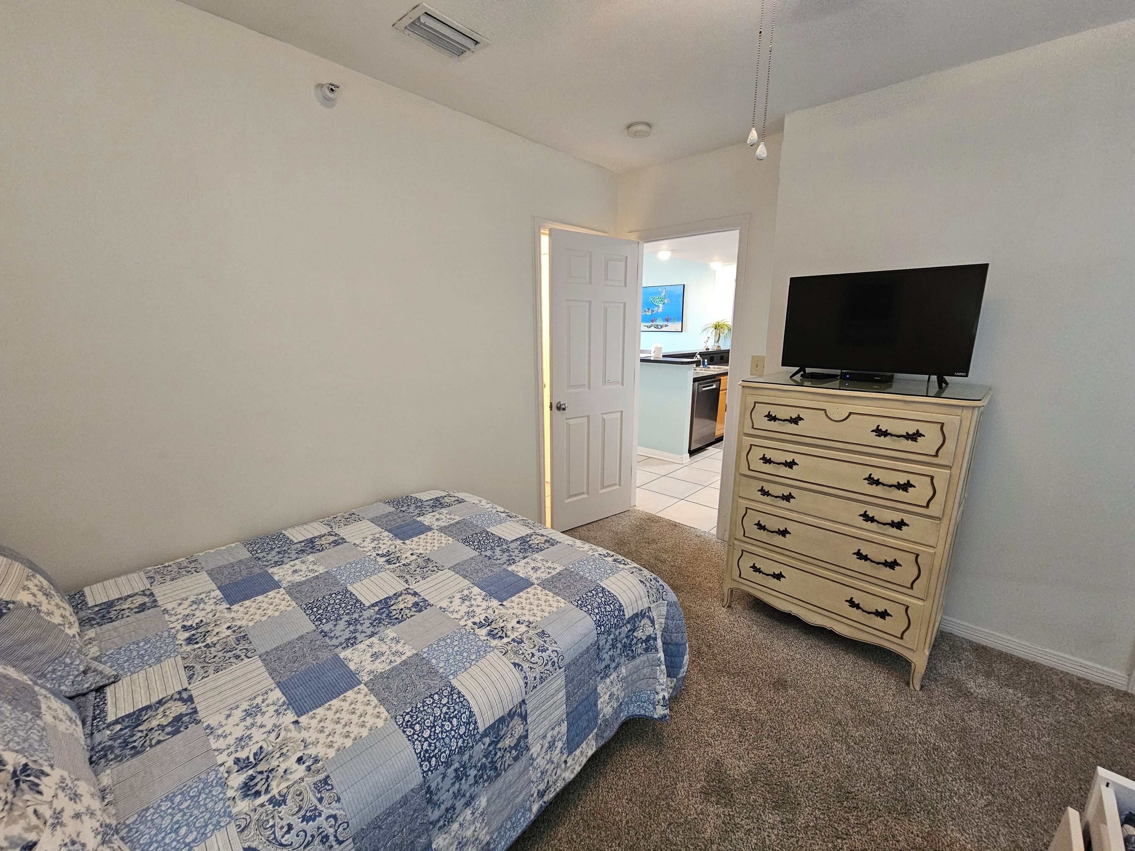 Une chambre à coucher comprenant un lit, une commode, un téléviseur et donnant sur la cuisine.