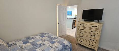 Une chambre à coucher comprenant un lit, une commode, un téléviseur et donnant sur la cuisine.