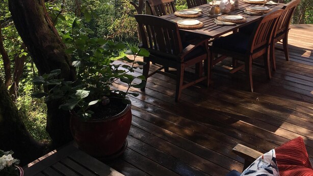 Una terraza de madera con mesa y sillas, rodeada de árboles y con vista al agua.