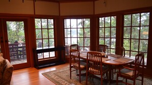 Un comedor con mesa y sillas de madera, amplias ventanas con vistas a árboles y suelo de madera.