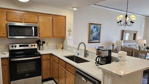 A modern kitchen with wooden cabinets, stainless steel appliances, a central island with a sink, and a dining area with a chandelier. - Inn at Dauphin Island 203 Beautifully Furnished with Great Views!! (Dauphin Island)