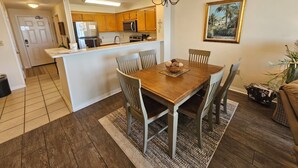 A dining area with a wooden table and chairs, a kitchen in the background, and a chandelier. - Inn at Dauphin Island 203 Beautifully Furnished with Great Views!! (Dauphin Island)