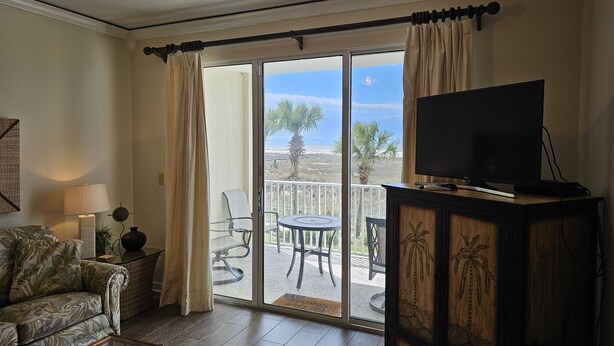 A living room with a sofa, a glass coffee table, a TV on a wooden cabinet, and a balcony with a view of palm trees and a clear sky. - Inn at Dauphin Island 203 Beautifully Furnished with Great Views!! (Dauphin Island)