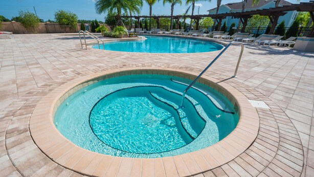 Uma banheira de hidromassagem circular com um deck revestido ao redor, uma piscina ao fundo, e cadeiras de descanso e guarda-sóis.