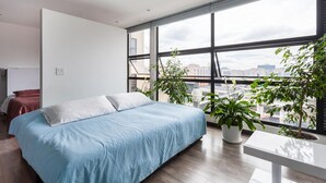 Un dormitorio moderno con una cama grande, vista de la ciudad a través de ventanales del piso al techo, y una planta en una mesita.