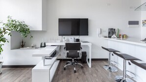 Un espacio de oficina moderno con un escritorio blanco, una silla de oficina negra, un televisor plano grande y una planta pequeña.