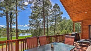 Terraza de madera con una mesa y sillas de vidrio, con vistas a un lago y un bosque.