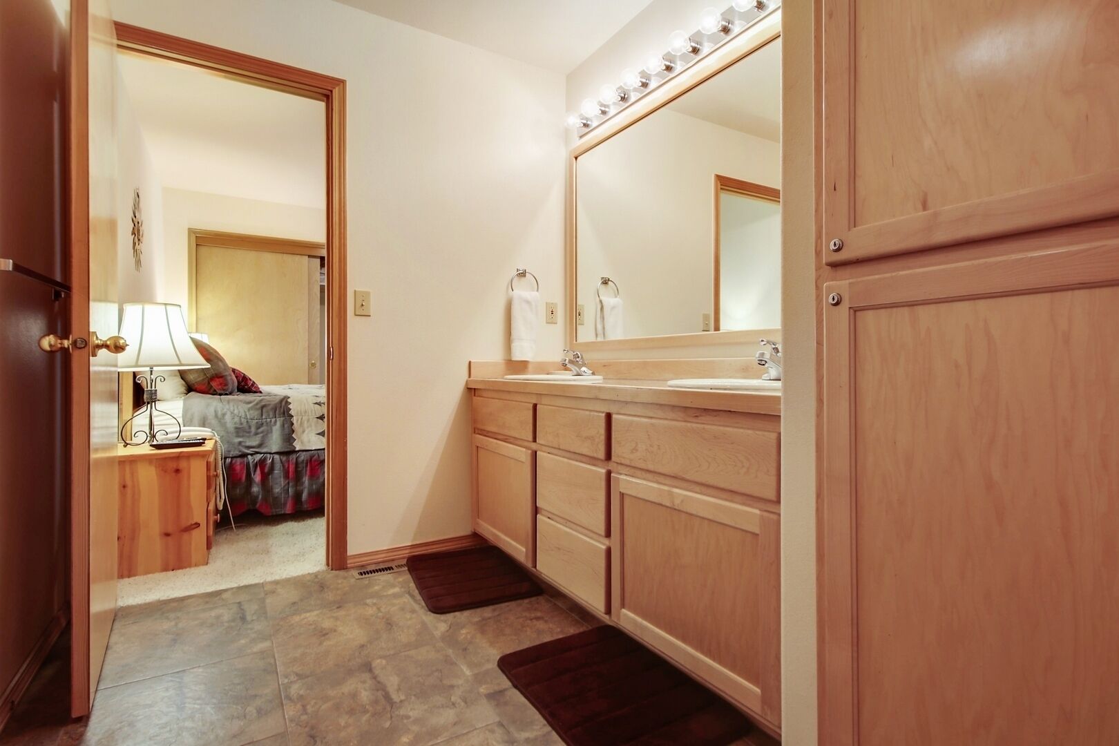 A bathroom with wooden cabinets, a large mirror with lights, a vanity with a sink, and a door leading to a bedroom.