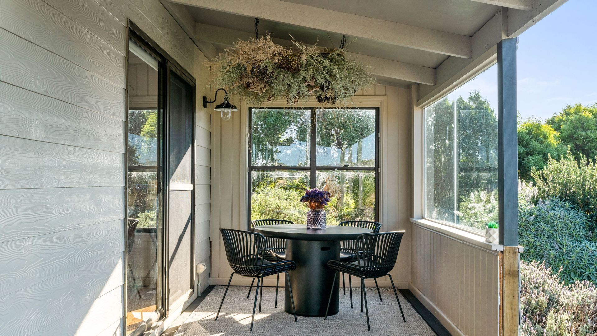 Une terrasse couverte avec une table et des chaises, de grandes fenêtres et des plantes suspendues.