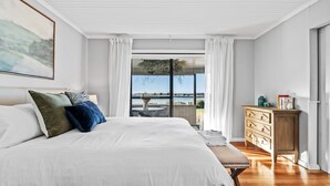Een slaapkamer met een groot bed, een houten dressoir en uitzicht op water door een glazen deur.