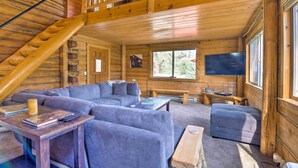 Interior de una cabaña de troncos con una escalera de madera, un sofá modular, un televisor de pantalla plana y un ventanal grande.
