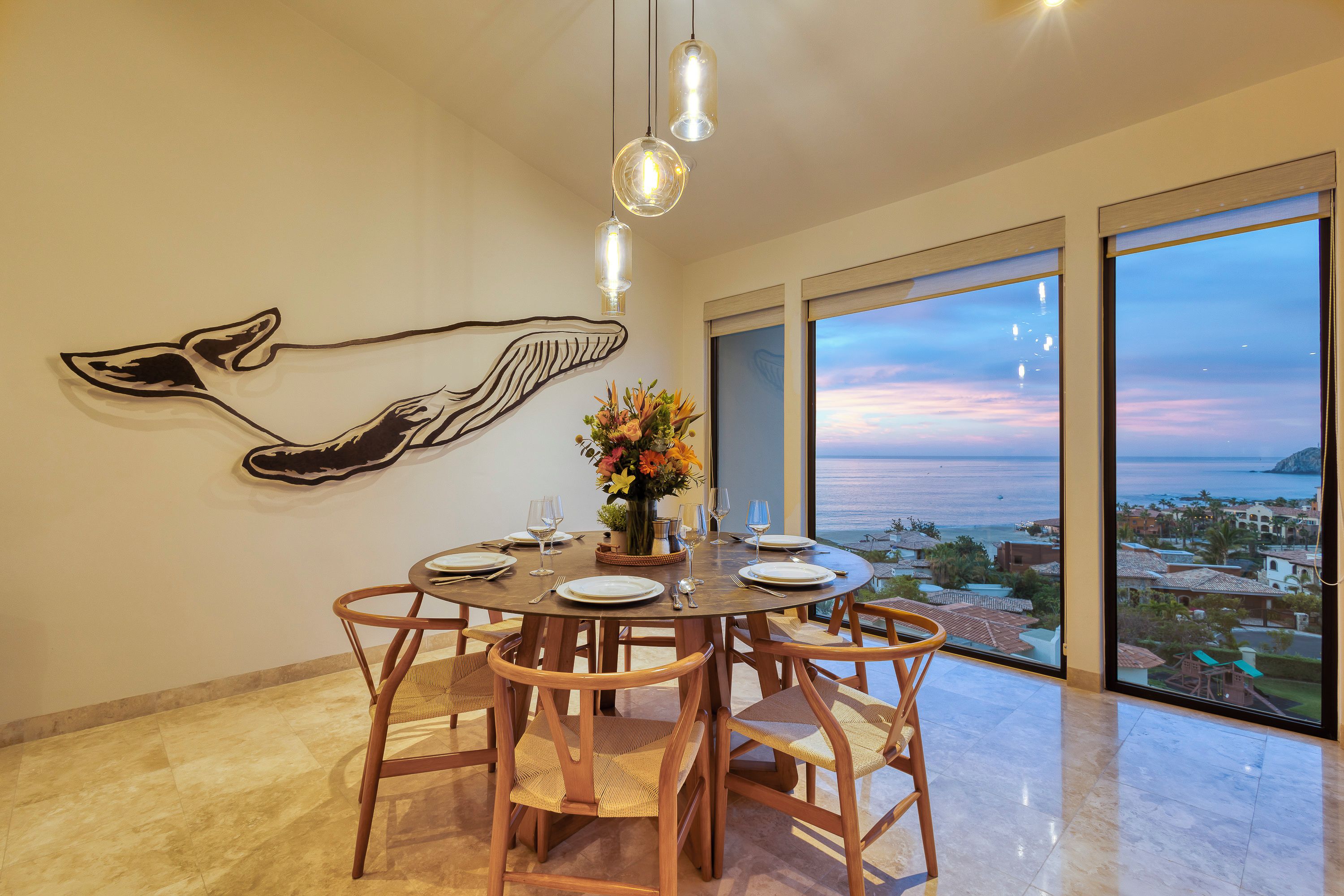 A dining area with a round table set for four, wooden chairs, a whale mural on the wall, and a view of the ocean through large windows.