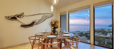 A dining area with a round table set for four, wooden chairs, a whale mural on the wall, and a view of the ocean through large windows.