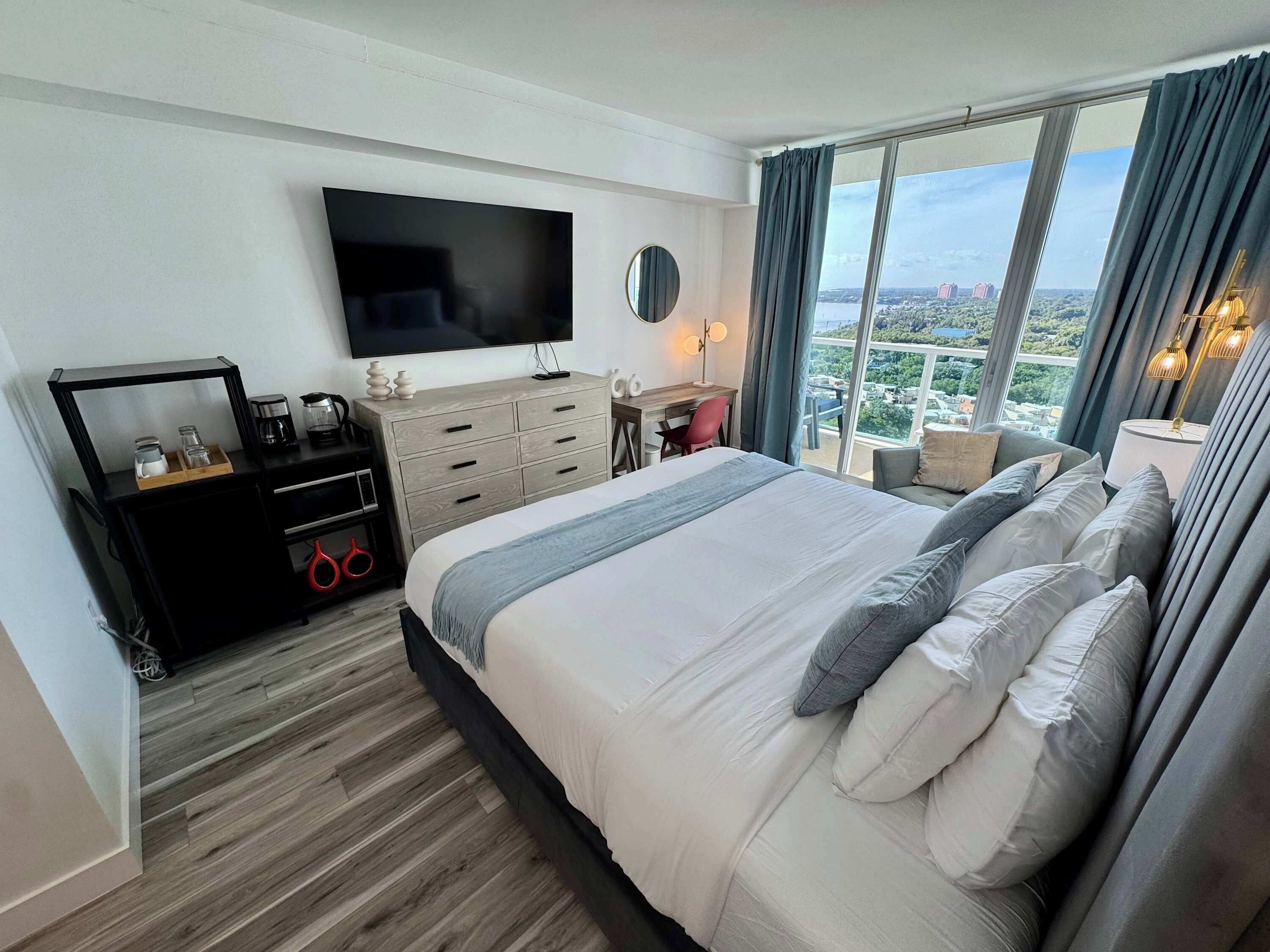 A hotel room with a large bed, a TV mounted on the wall, a desk with a chair, and a view of the city through large windows.