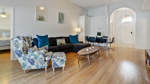 A modern living room with a dark-colored sofa, patterned armchairs, a wooden coffee table, and a dining area with a glass table and chairs.