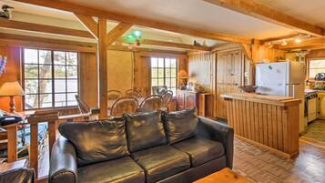 A cozy cabin interior with a black leather sofa, wooden dining table, chairs, and a kitchen area with a refrigerator and counter.