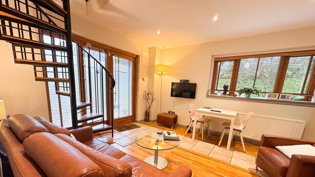 A living room with a brown leather sofa, a glass coffee table, a white dining table with two chairs, a TV on the wall, and a staircase with a wooden railing. - The Lodge at Bodenlodge is located in the charming Costal Village of Malahide (Malahide)