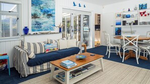 Ein Wohnzimmer mit einer Couch, einem Couchtisch und einem Essbereich mit Stühlen. Ein Bücherregal mit verschiedenen Gegenständen und ein Gemälde an der Wand.