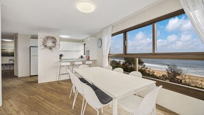 A modern kitchen with white cabinets, a dining area with a long table and white chairs, and a large window overlooking the beach. - Parkyn Place 6 - Three Bedroom Oceanview Apartment on Mooloolaba Spit (Mooloolaba)