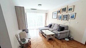 A modern living room with a grey sofa, a white coffee table, and a floor lamp. There are framed pictures on the wall and a window with curtains. - Apartman MAJDA with balkony (Split)