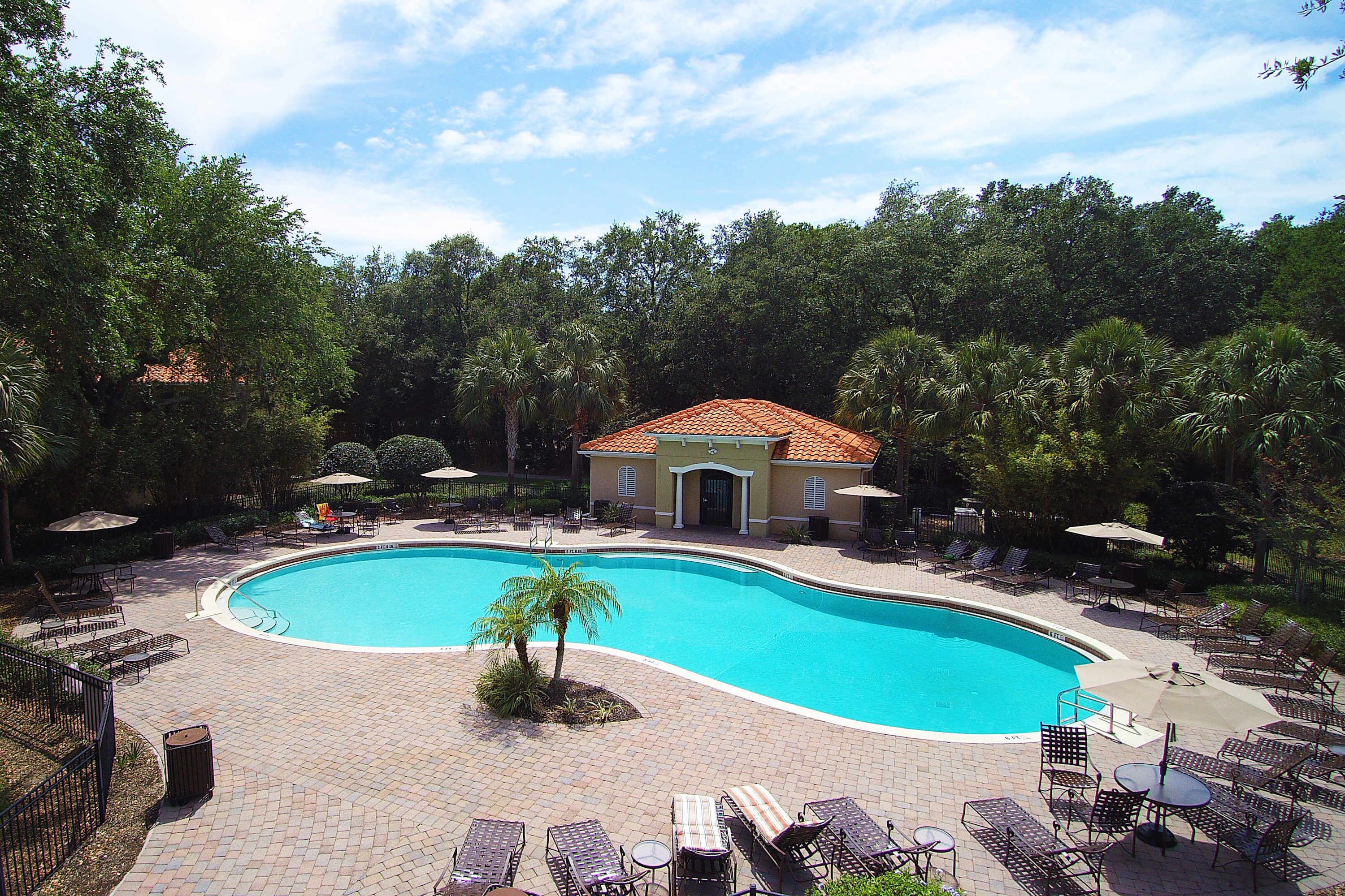 A swimming pool with lounge chairs and umbrellas, surrounded by trees and a building with a tiled roof.