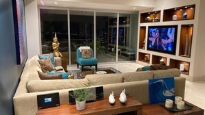 Uma sala de estar moderna com um sofá grande, uma mesa de centro e uma televisão. Há uma janela ampla com vista para a cidade à noite.