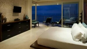 Quarto com cama grande, televisão fixada na parede, varanda com vista para a cidade à noite e ventoinha no teto.