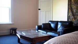 Una habitación con un sofá de cuero negro, una mesita de centro de madera, un taburete pequeño y una cama con colcha estampada.