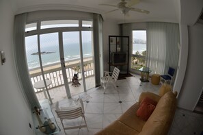 Una sala de estar con un gran ventanal que da a un balcón con vista al océano y la playa.