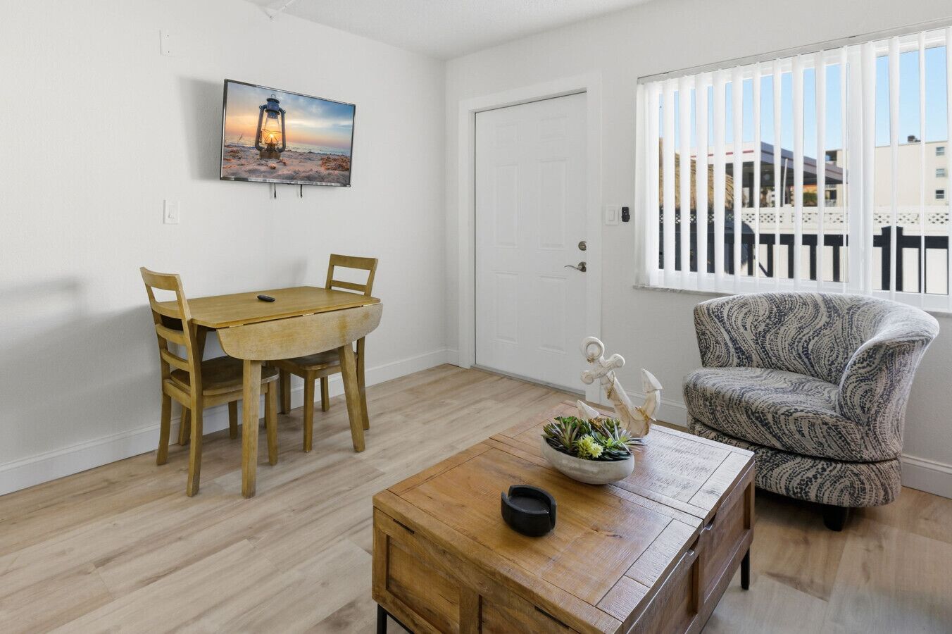 A room with a wooden dining table, two chairs, a sofa, a coffee table with a plant, a door, and a window with blinds.