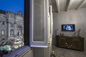 Un interior con vista a una fuente histórica exterior, un mueble de madera con objetos decorativos y un televisor en la pared.