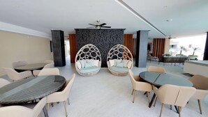 Un área de estar interior moderna con mesas y sillas redondas, dos sillones colgantes tipo huevo y un ventilador de techo.