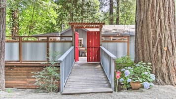 Une terrasse en bois menant à une porte rouge, entourée d’une clôture en bois et d’un grand arbre.