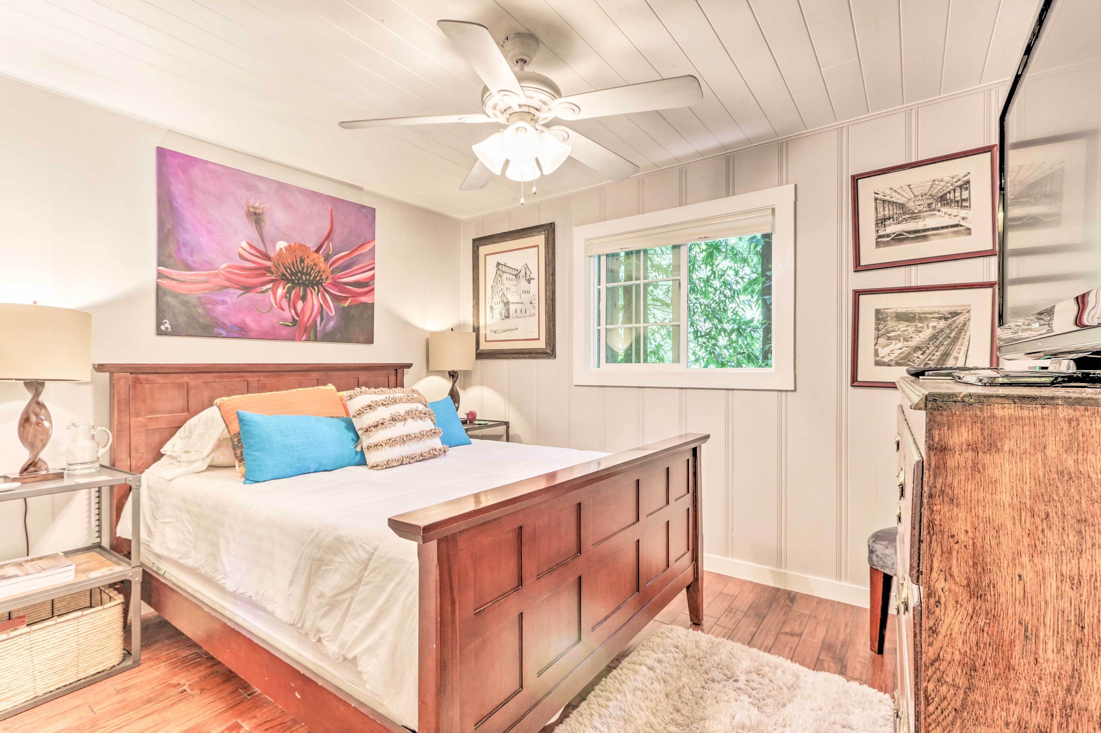 Une chambre avec un lit en bois, un ventilateur de plafond, une fenêtre donnant sur un paysage verdoyant et des tableaux encadrés accrochés au mur.