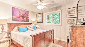 Une chambre avec un lit en bois, un ventilateur de plafond, une fenêtre donnant sur un paysage verdoyant et des tableaux encadrés accrochés au mur.