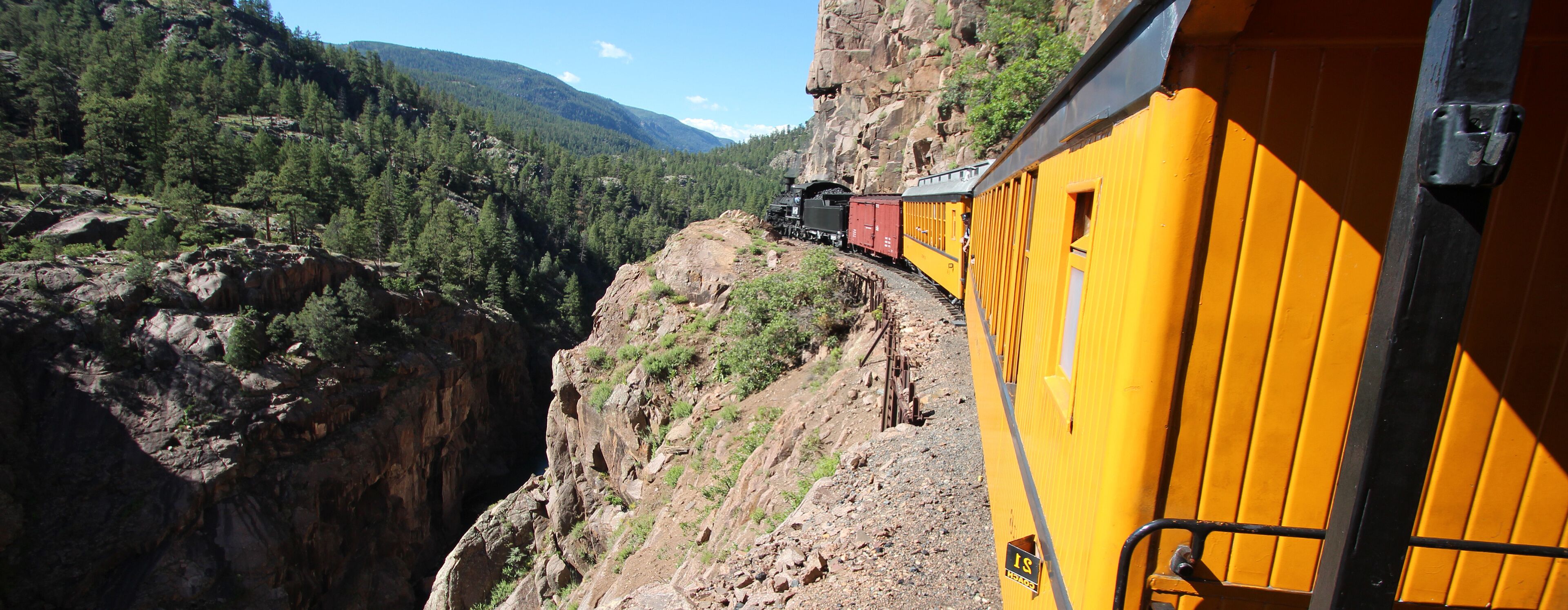 A train on a mountain track with a yellow train car on the right.