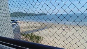 Vista de la playa a través de una cerca de alambre, con un edificio y palmeras en primer plano.