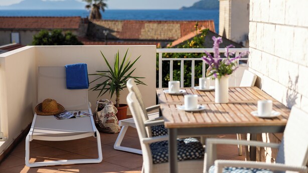 Ein Balkon mit Holztisch, weißen Stühlen, einer Liege, einem Topfpflanzen und Blick auf das Meer und die Berge.