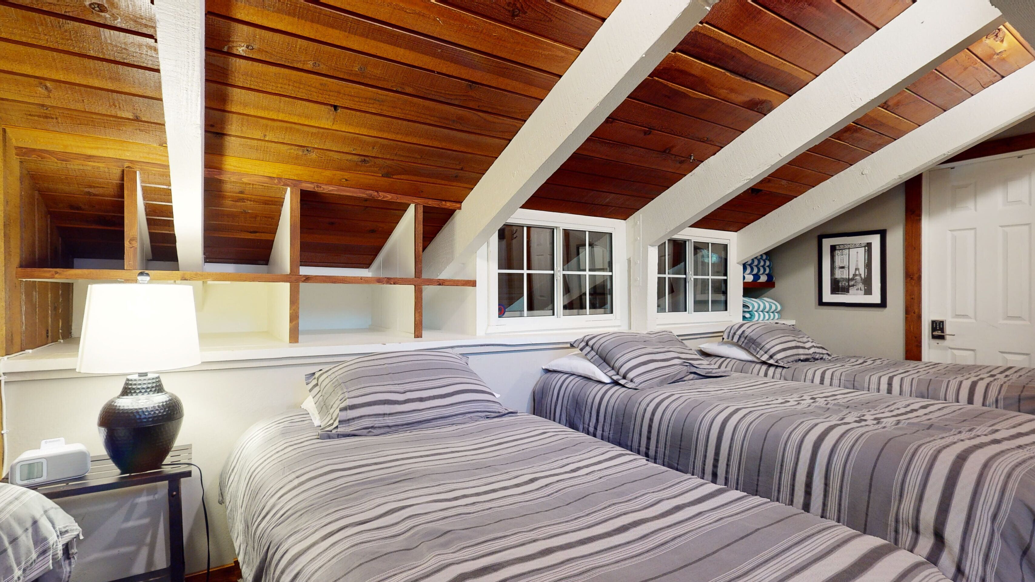 A bedroom with two beds, wooden ceiling beams, a window, and a bedside table with a lamp.